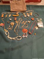 Overview of assorted vintage women's jewelry laid out on a dark green fabric, showing necklaces, earrings, rings, brooches, and bracelets including a pumpkin brooch and white beaded neckwear.