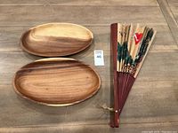 Photo showing two oval shallow carved wooden trays and one folding hand fan placed on a wooden surface.