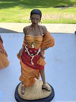 Figurine of a woman standing with orange shawl, red skirt, shell jewelry, hoop earrings, and white sandals on a wooden base outdoors.