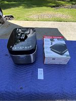 Photo of an electric pressure cooker and boxed George Foreman indoor grill on a blue moving blanket outside on concrete.
