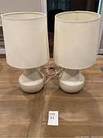 Pair of off white table lamps with matching off white fabric shades, placed side by side on wooden floor with cords visible.