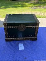 Photo of a small rectangular trunk with dark covering and metal studs and clasp, placed on a table with a numbered card in front.