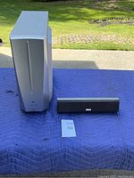 JVC powered subwoofer and center channel speaker shown on outdoor surface. Subwoofer is tall silver rectangle. Center channel speaker is smaller, black and rectangular.