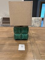 Photo of square turquoise ceramic table lamp with beige square fabric lampshade on wooden table.