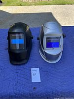 Two welding helmets side by side on a blue padded mat outside on pavement with grass in background.