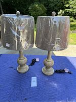 Photo shows two turned wooden table lamps with new gray fabric shades covered in plastic. Both lamps have electrical cords attached.