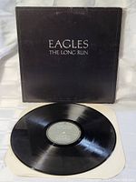 Photo of the black vinyl record LP with original gray center label on white inner sleeve, and front of black album cover with white text Eagles The Long Run