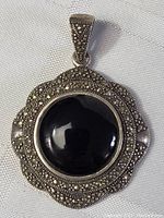Close-up image of large black Gothic sterling silver pendant with marcasite detailing and polished black central stone, displayed on white fabric background.