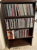 Two photos showing wooden shelving units with approximately 90-100 assorted music CDs arranged on two full shelves and a partial third shelf. CDs are standard jewel cases with mixed genres like adult contemporary, jazz, classic singers, and classical music.