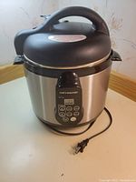 Front view of Cooks Essentials electric pressure cooker showing stainless steel body, black handle, and digital control buttons.