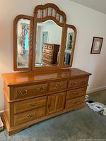 Front view of wood dresser showing three mirrors with foldable side leaves. Nine drawers in two rows below mirrors. Warm wood finish and brass hardware.