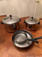 Photo showing Farberware cookware set on a wooden table, includes large stock pot, medium soup pot, two sauté pans (one smaller, one larger), all stainless steel with black handles and matching stainless steel lids with black knobs.