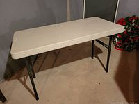 Photo showing rectangular cream-colored plastic craft table with black metal foldable legs.