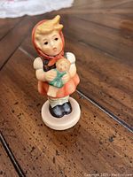 Front view of Goebel figurine showing girl holding doll, detailed hand-painted features, and red hood.