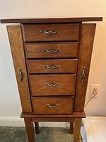 Front view of wooden jewelry box showing five drawers with ornate metal handles and two side doors with matching handles on a stand.