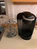 Black Keurig coffee maker with water reservoir on the left, accompanied by a circular metal spinning K-Cup pod holder on a wooden surface, against a floral wallpaper background.