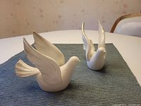 Two white porcelain dove figurines displayed on a blue cloth, showing sculpted wings and tails, in daylight.