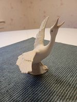 Side view of the white matte finish porcelain flying goose figurine on a textured fabric surface, showing detailed sculpted feathers and open beak.
