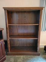 Empty dark walnut bookshelf with three shelves and decorative carved top edge.