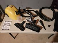 Photo of Steam Buggy main unit on a white table with all attachments including brushes, nozzles, a hose, extension rod, measuring cup, and instruction manual.