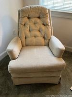 Beige swivel rocker chair positioned near a window showing tufted backrest with button details, padded armrests, and reversible seat cushion with visible texture of brushed velvet upholstery.