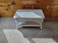 White wicker coffee table positioned in front of a wooden paneled wall on carpeted flooring, showing the entire table including the top surface and lower shelf.