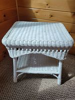 White wicker side table with square top and lower shelf against wooden wall and carpeted floor