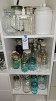 Three shelves filled with multiple Ball mason jars in clear and blue glass, glass bottles with stoppers, and two white ceramic storage canisters with lids.