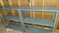 Two gray low metal shelves with three shelves each, showing minor wear and situated in an unfinished room with wood wall and concrete floor.