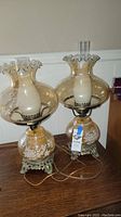 Two vintage hurricane lamps with clear glass globes, ruffled tops, floral decoration, and metal bases on a wooden surface.