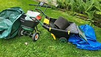 Photo of outdoor garden equipment including a green broadcast seed spreader with cloth collection bag, a green Miracle-Gro wheeled cart, stacked black plastic plant pots, and blue tarps on grass.