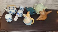 Photo showing the collection of ceramic teapots, matching cups, and animal figurines arranged on a dark surface against a light curtain backdrop.