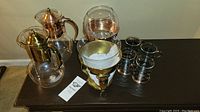 Overview of vintage copper and glass coffee set on dark wood table, including carafe, warming stand, glass cups with metal holders, and instruction manual.