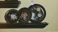 Close-up of three round trays on a shelf showing hand-painted pink floral designs on black toleware with gold accents.
