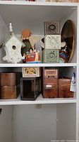 Shelf displaying wooden drawers, decorative wooden blocks, figurines, a white birdhouse, a dark wooden crate, a vanilla tea metal box, and a large brass tray visible on the side.