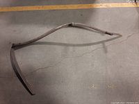 Photo showing one curved wooden scythe handle without blades, lying on concrete floor.