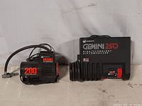 Two mini air compressors on a flat surface. Left is the Mini 200 air compressor, right is the Gemini 250 air compressor with coiled hose.