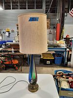 Front view of the lamp showing the glass body, brass base and beige cylindrical fabric lampshade with some marks or wear on fabric