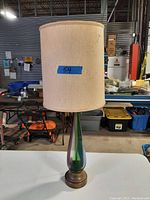 Front angle view of the lamp shows the tall beige drum shade and iridescent blown glass base on a white table in front of workshop tools and storage bins.