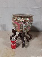 Full view of a round Chinese porcelain planter with colorful Chinoiserie paintings, sitting on a dark wooden stand with carved legs. A soda can next to it shows scale, indicating about 8 inches height and 10 inches top diameter.