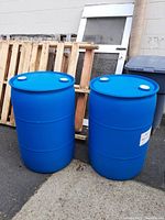 Two upright blue plastic rain barrels next to a wall and wooden pallets.
