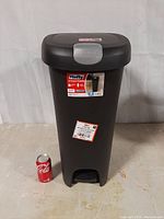 Front view of Hefty 12 gallon black plastic step-on trash can with label visible and a Coca-Cola can for size reference.