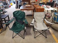 Two folding camp chairs side by side in warehouse setting, one green, one light gray, with cup holders in armrests.