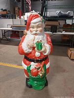 Front view of the large vintage blow mold Santa holding a green gift and pointing, standing on a green sack with toys.