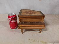 Front and side view of vintage wooden jewelry music box in the shape of a piano with floral artwork on lid and painted piano keys on front next to a soda can for scale.