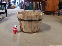 Wooden bucket planter shown next to a Coca-Cola can for scale. The planter is approximately 12 inches tall and 18 inches across the top. Light wood with two black metal bands and a metal bottom rim.