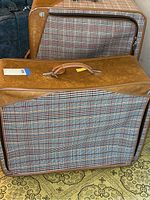 Front angled view of two plaid canvas sided luggage suitcases with brown trim stacked together. Shows overall shape, design, and handle of luggage.
