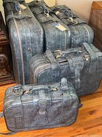 Five matching blue leather luggage cases arranged from largest to smallest. The cases have matching straps with buckle closures and handles. Four cases have wheels visible at bottom edges.