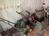 Full view of four older gas mowers and a red gas roto tiller inside a storage shed, showing green vintage mowers and handles.