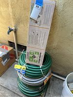 Four coiled garden hoses stacked along a wall with a boxed lawn watering sprinkler on top.
